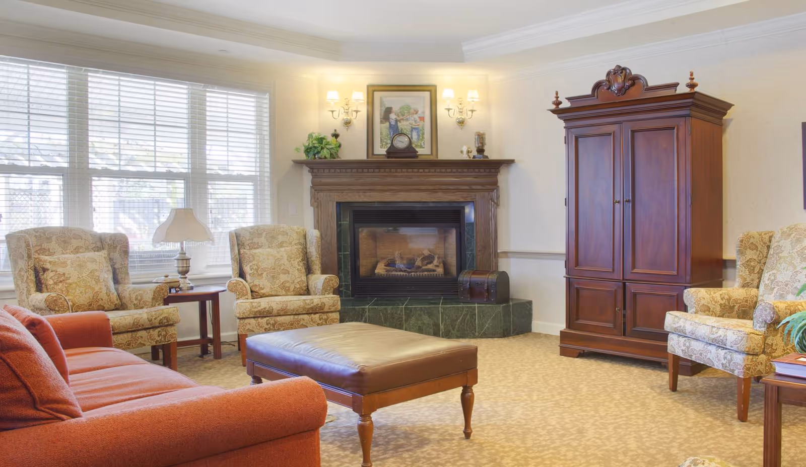 A cozy living room with a red sofa, two patterned armchairs, a large ottoman, a wooden armoire, and a fireplace with a mantle decorated with a clock, a framed picture, and small plants. Large windows with white blinds allow natural light to fill the room.