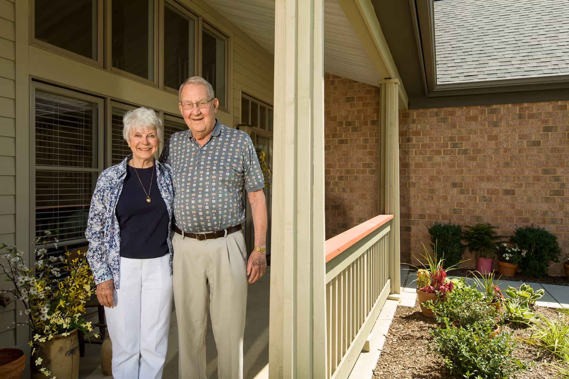 An elderly couple standing together and smiling on a porch outside a building with beige siding and large windows. There are potted plants and flowers around them, and a garden area with shrubs and flowers is visible to the right.