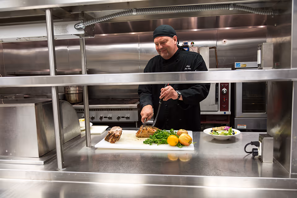 A chef wearing a black uniform and head covering is slicing cooked meat on a cutting board in a commercial kitchen. Fresh vegetables and a prepared salad plate are also visible on the counter.