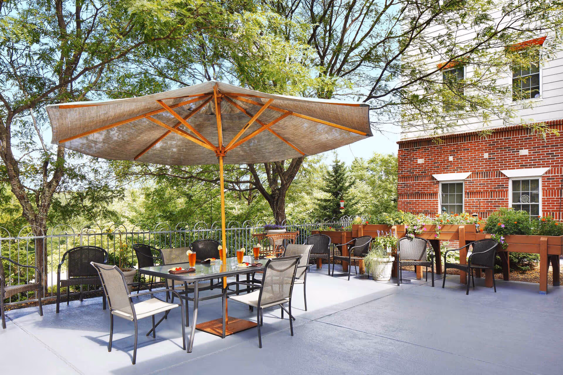 Outdoor patio with a large umbrella over a dining table and chairs, potted plants, and a brick building in the background.