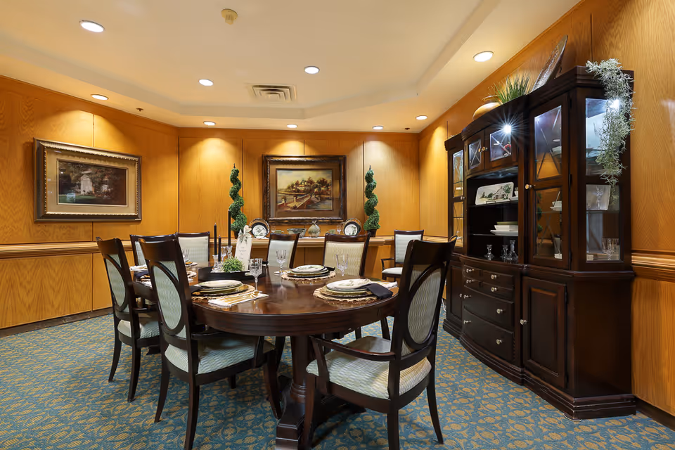 A formal dining room with a large oval wooden table set with plates, glasses, and napkins. Eight upholstered chairs surround the table. The room has wood-paneled walls, two framed paintings, two decorative topiary plants, and a dark wooden china cabinet displaying dishes and glassware. The carpet is patterned in blue and gold tones, and recessed ceiling lights illuminate the space.