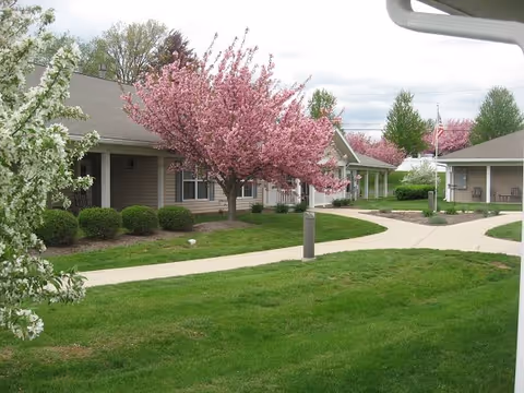 A landscaped outdoor area of a senior living community with green grass, paved walkways, and blooming pink and white flowering trees. Single-story buildings with beige siding and gray roofs are visible in the background under a cloudy sky.