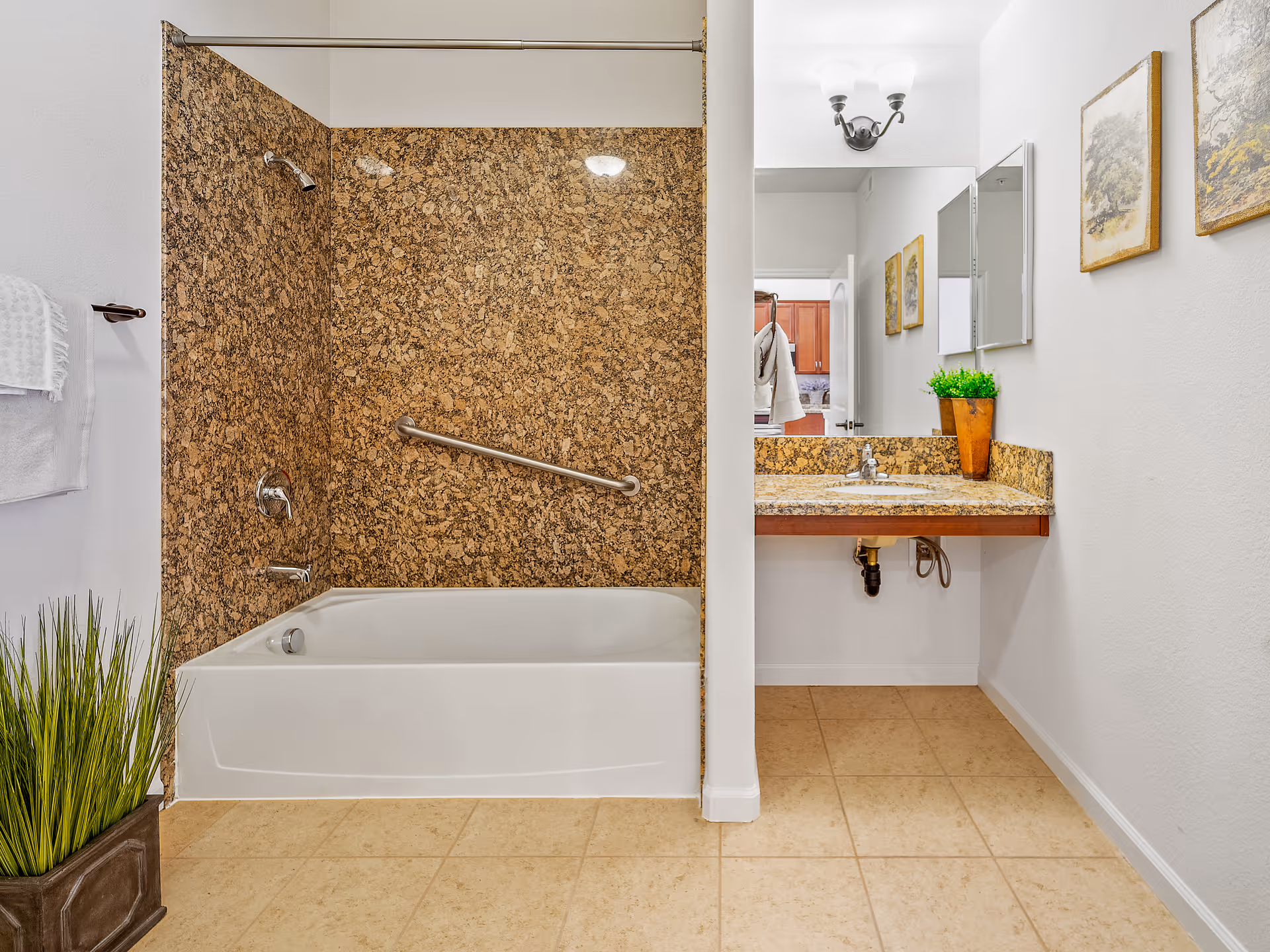 A clean bathroom with a white bathtub and brown granite wall surround. There is a metal grab bar on the wall above the bathtub. To the right, there is a sink with a brown granite countertop and a mirror above it. The floor is tiled, and there are two framed pictures on the white wall next to the sink. A potted plant is placed on the floor near the bathtub, and another plant is on the countertop.