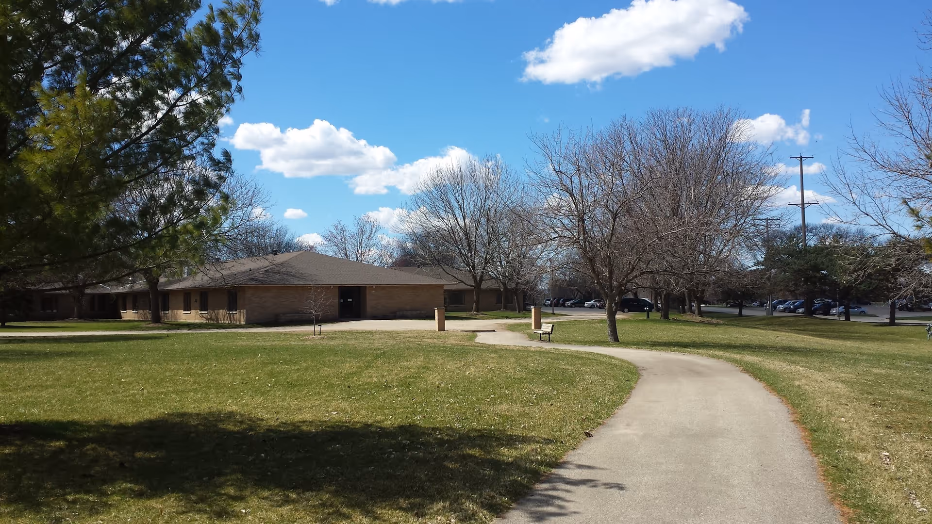 A paved walking path curves through a grassy area with leafless trees and a bench. In the background, there is a single-story brick building under a partly cloudy blue sky. Several parked cars are visible beyond the trees.
