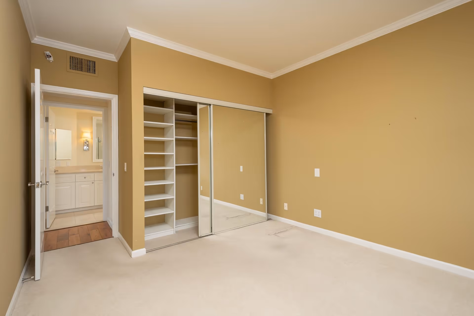 Empty bedroom with beige walls and carpet, featuring a closet with sliding mirrored doors and built-in shelves. An open door leads to a bathroom with white cabinetry and a light fixture above the mirror.