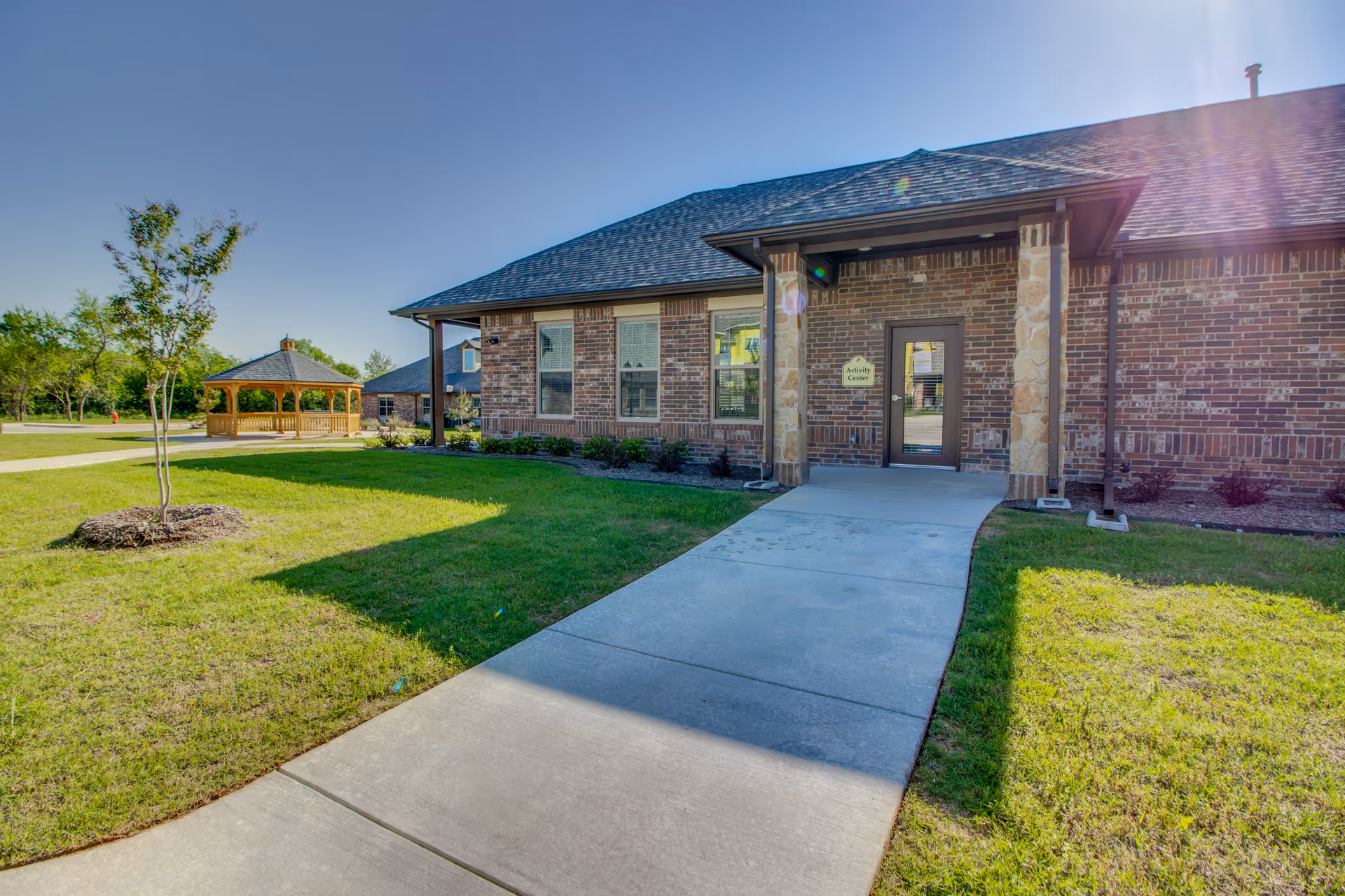 Brick exterior entrance of an assisted living building with a concrete walkway, lawn, and a wooden gazebo to the left.