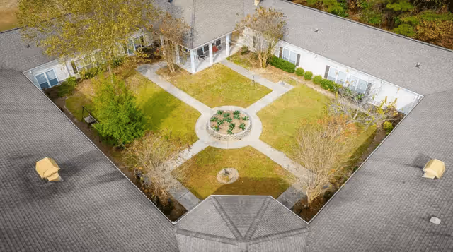 Aerial view of a rectangular courtyard garden surrounded by a single-story building with a gray roof. The courtyard features a circular flower bed in the center with pathways extending outward in four directions, bordered by grass and small trees.