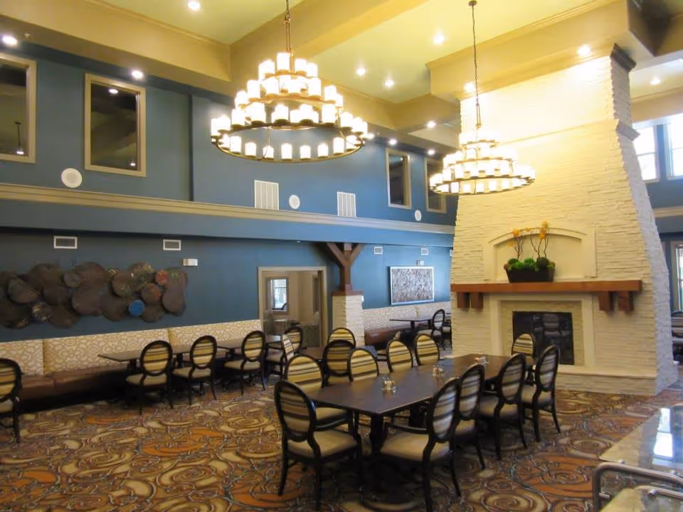A spacious dining room with a large rectangular table surrounded by chairs with striped upholstery. The room features a tall white stone fireplace with a wooden mantel, decorated with a plant. Two large circular chandeliers hang from the high ceiling. The walls are painted blue with decorative elements and windows near the ceiling. The floor has a patterned carpet.