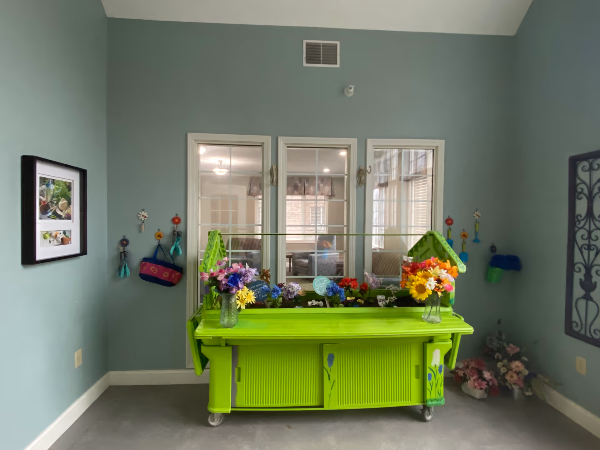 A bright green gardening cart filled with colorful artificial flowers is placed against a light blue wall. Behind the cart are three white-framed windows looking into another room. On the left wall, there is a framed picture and several hanging gardening tools and gloves. On the right side, there is a decorative black metal wall hanging and more artificial flowers in a corner.