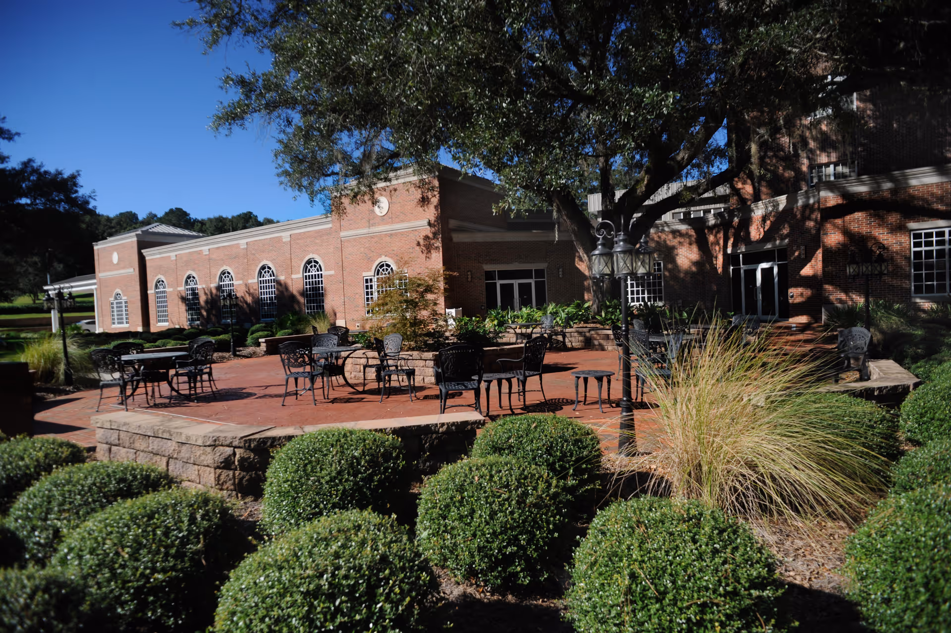 Brick senior living building with a shaded outdoor patio area featuring metal tables and chairs and manicured shrubs.
