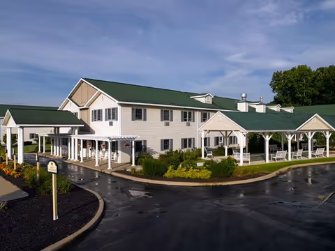 Exterior view of a two-story beige senior living facility with green roofs, covered entrances, walkways, and landscaped grounds.