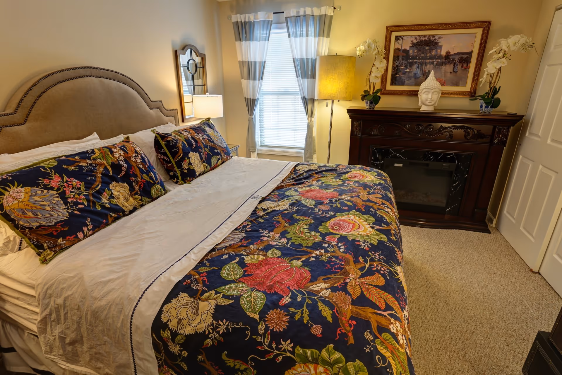 A cozy bedroom featuring a large bed with a beige upholstered headboard and colorful floral bedding. The room has a window with white and gray striped curtains, a bedside table with a lamp, and a dark wooden electric fireplace mantel decorated with white orchids and a Buddha head sculpture. A framed painting hangs above the fireplace.