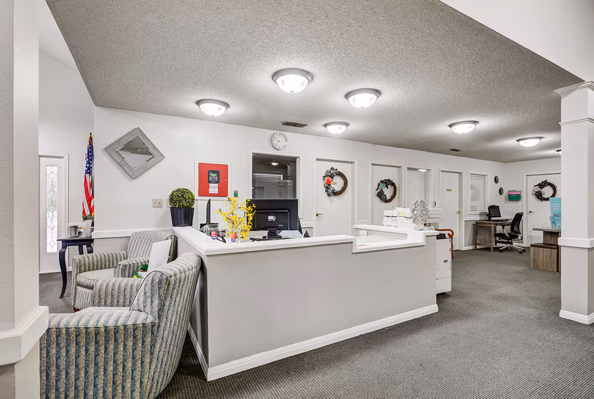 Reception desk and seating area in a well-lit senior living facility lobby with chairs, workstations, and wall wreaths.