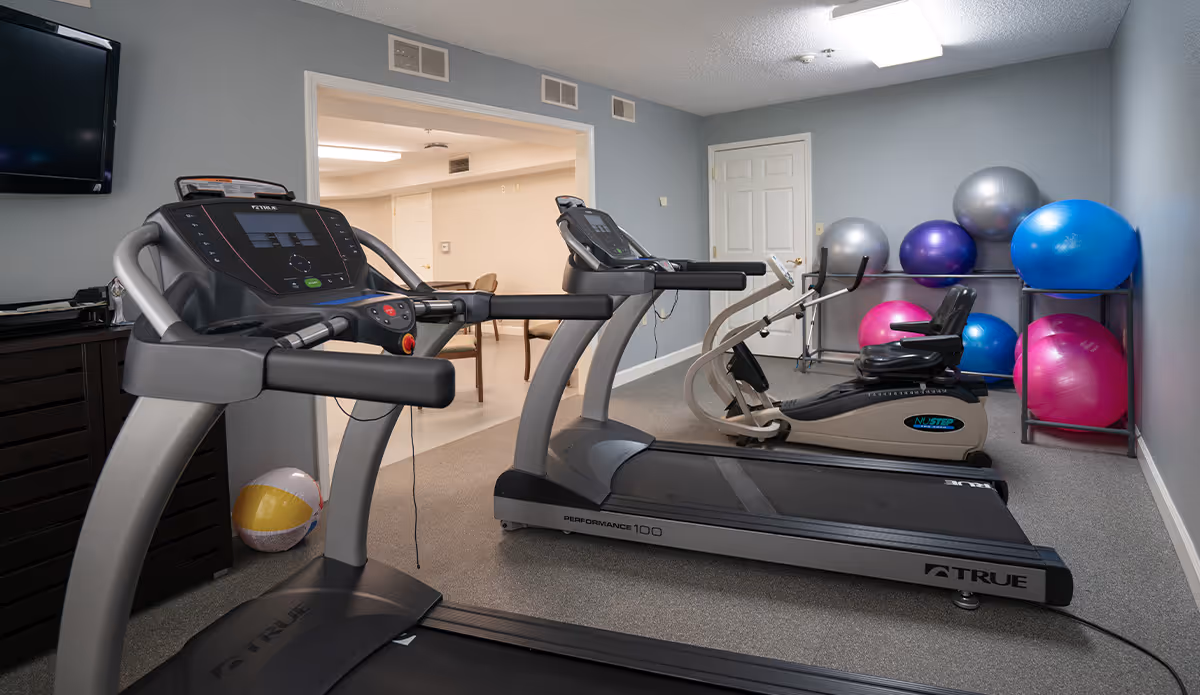 Indoor fitness room with two treadmills, a recumbent exercise bike, and a rack holding several large exercise balls in various colors including silver, purple, blue, and pink. A television is mounted on the wall to the left, and a doorway leads to another room with tables and chairs.