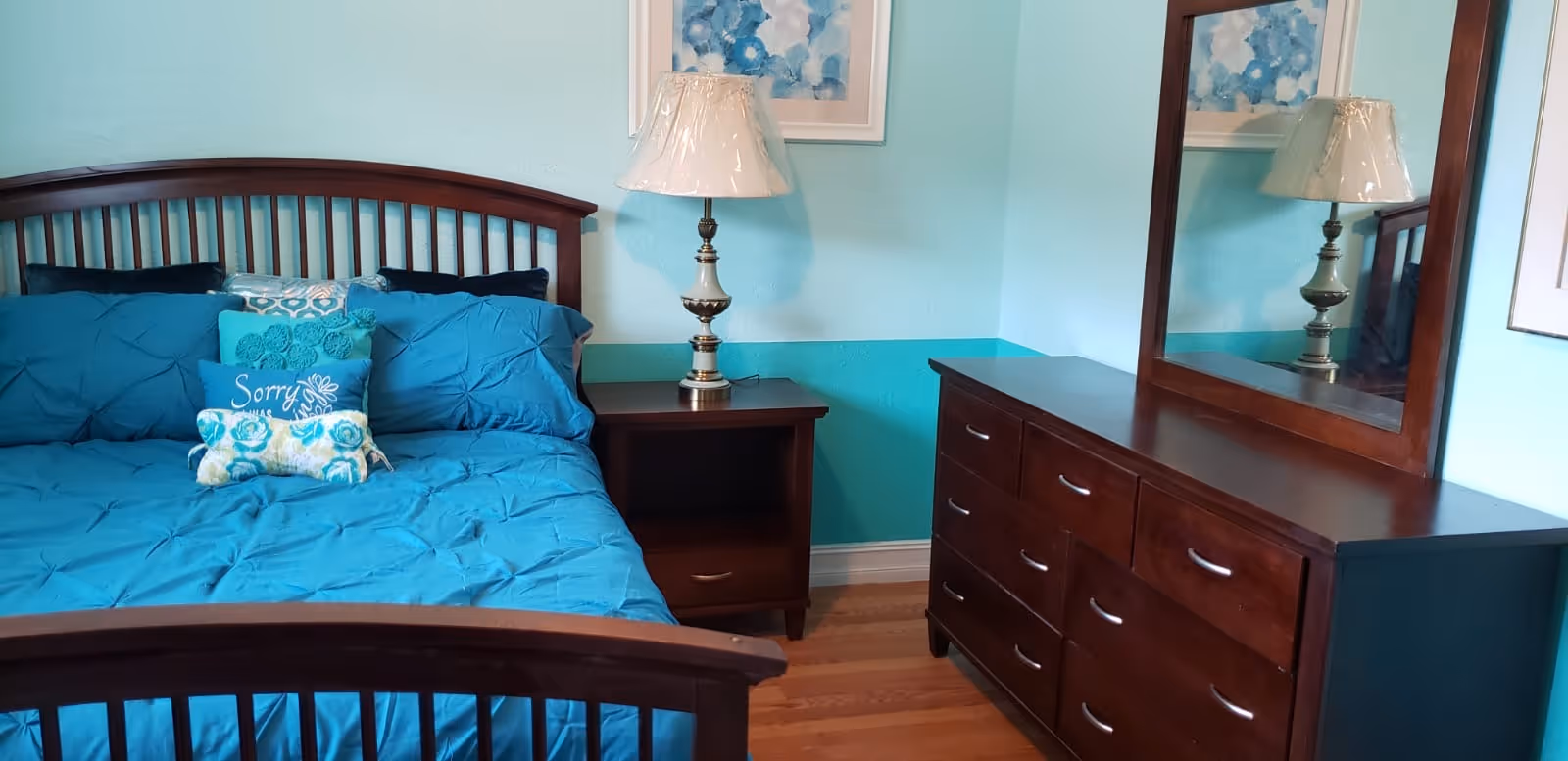 Bedroom with a wooden bed and teal bedding, matching pillows, a nightstand with lamp, and a wooden dresser with mirror.