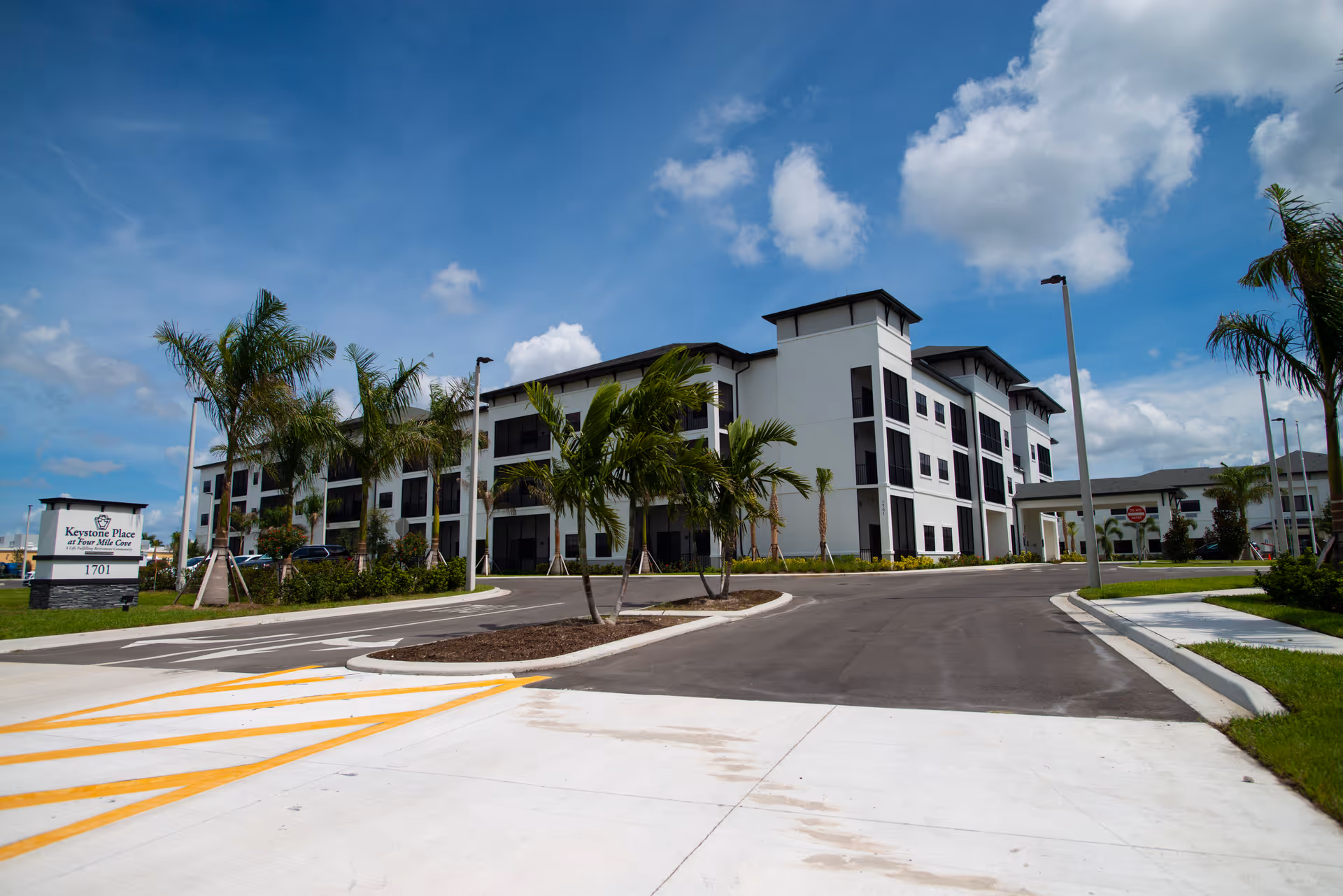 Exterior view of a modern three-story senior living facility building with palm trees and a clear blue sky. The driveway and parking area are visible in the foreground, along with a sign that reads 'Keystone Place at Tara Mills Cove 1701'.