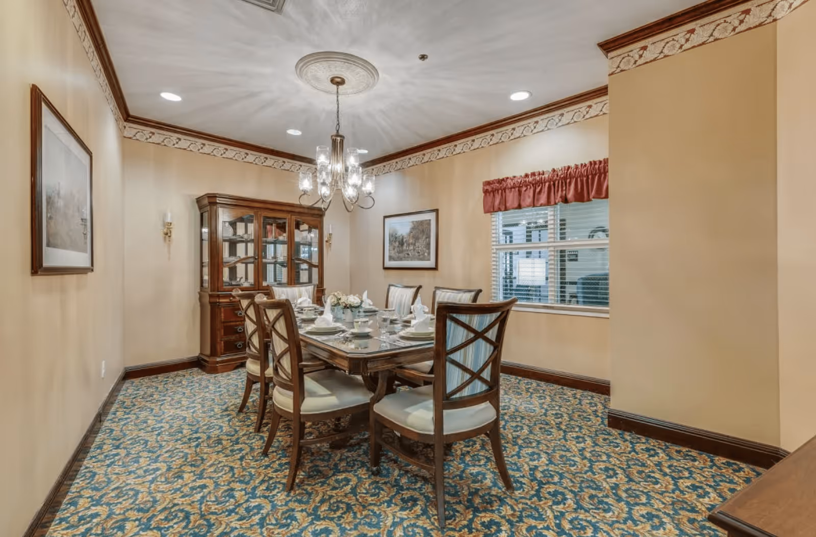 A formal dining room with a rectangular wooden dining table set for six people. The chairs have cushioned seats and wooden backs with a crisscross design. A chandelier with multiple lights hangs above the table. There is a wooden china cabinet with glass doors against the far wall, and two framed pictures hang on the walls. The room has beige walls with a decorative border near the ceiling, a patterned carpet in blue and gold tones, and a window with blinds and a red valance.
