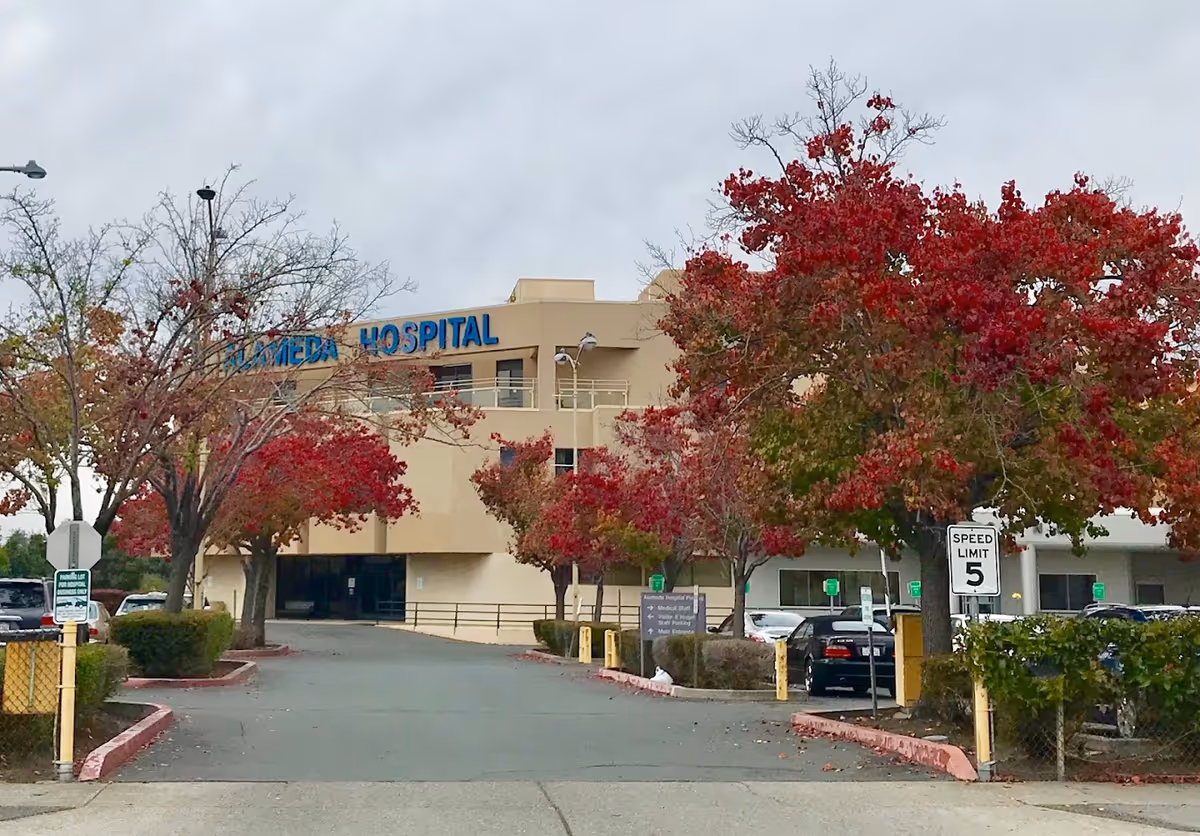 Exterior view of Alameda Hospital building with beige walls and blue signage. The scene includes a driveway leading to the entrance, surrounded by trees with red and green autumn leaves, parked cars, and a speed limit sign of 5 mph.