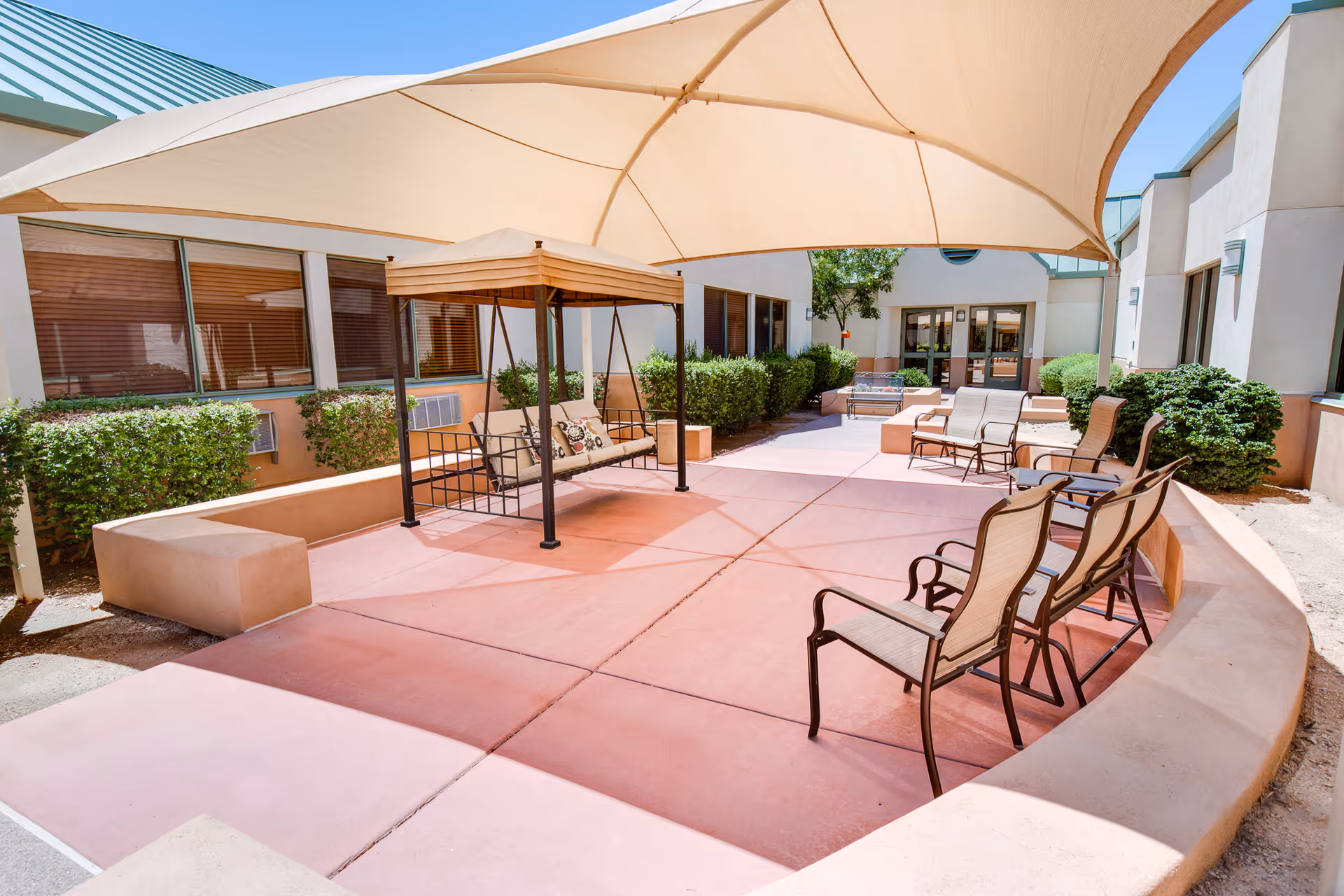 Outdoor courtyard area with a large beige canopy providing shade over a swing bench and several patio chairs arranged on a reddish concrete floor. The courtyard is surrounded by building walls with windows and green bushes, and there is a tree visible in the background near the entrance doors.