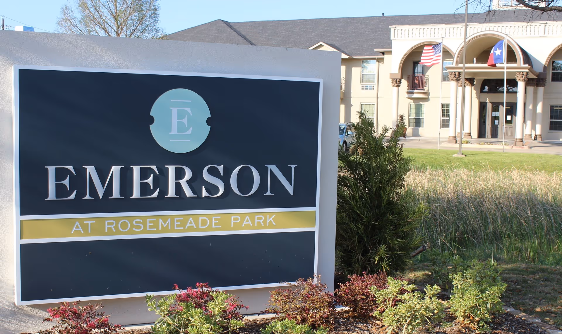 Outdoor view of the entrance to Emerson at Rosemeade Park facility with a large sign displaying the facility name in front, landscaping with bushes and grass, and a building with columns and flags in the background.