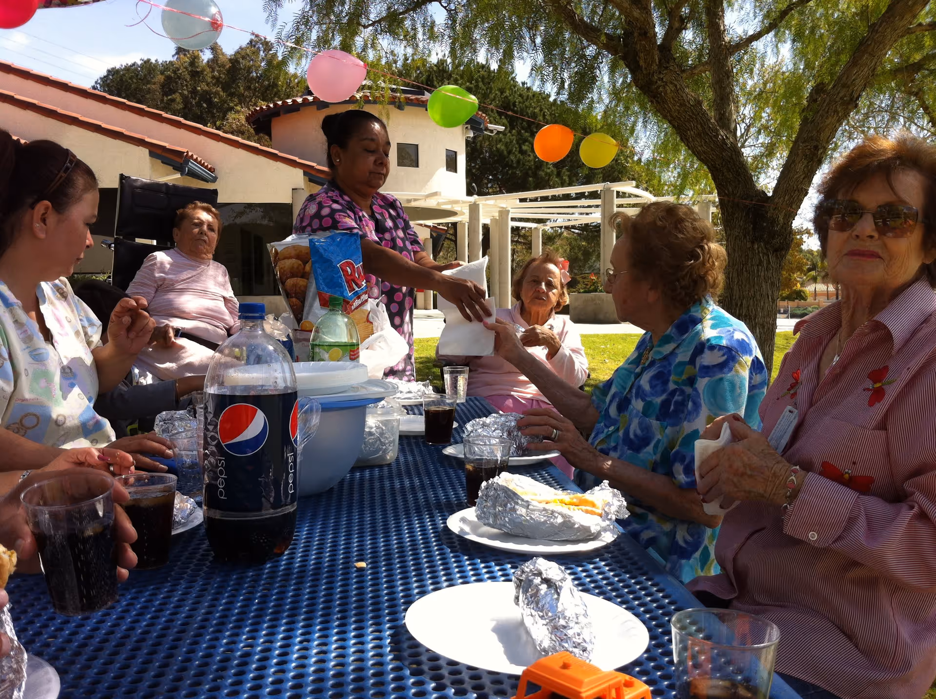 A group of elderly women and a caregiver sitting around a blue outdoor picnic table enjoying a meal together. The table has plates with food wrapped in foil, cups with dark beverages, a large bottle of Pepsi, and other food items. Colorful balloons are strung above them, and there is a tree and a building in the background.