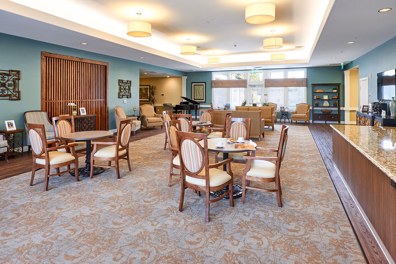 A spacious and well-lit common area in a senior living facility featuring multiple round tables with wooden chairs upholstered in cream and striped fabric. The room has a patterned carpet, teal walls, and several comfortable armchairs and sofas arranged near large windows. A grand piano is visible in the background, along with decorative wall art and a granite countertop on the right side.