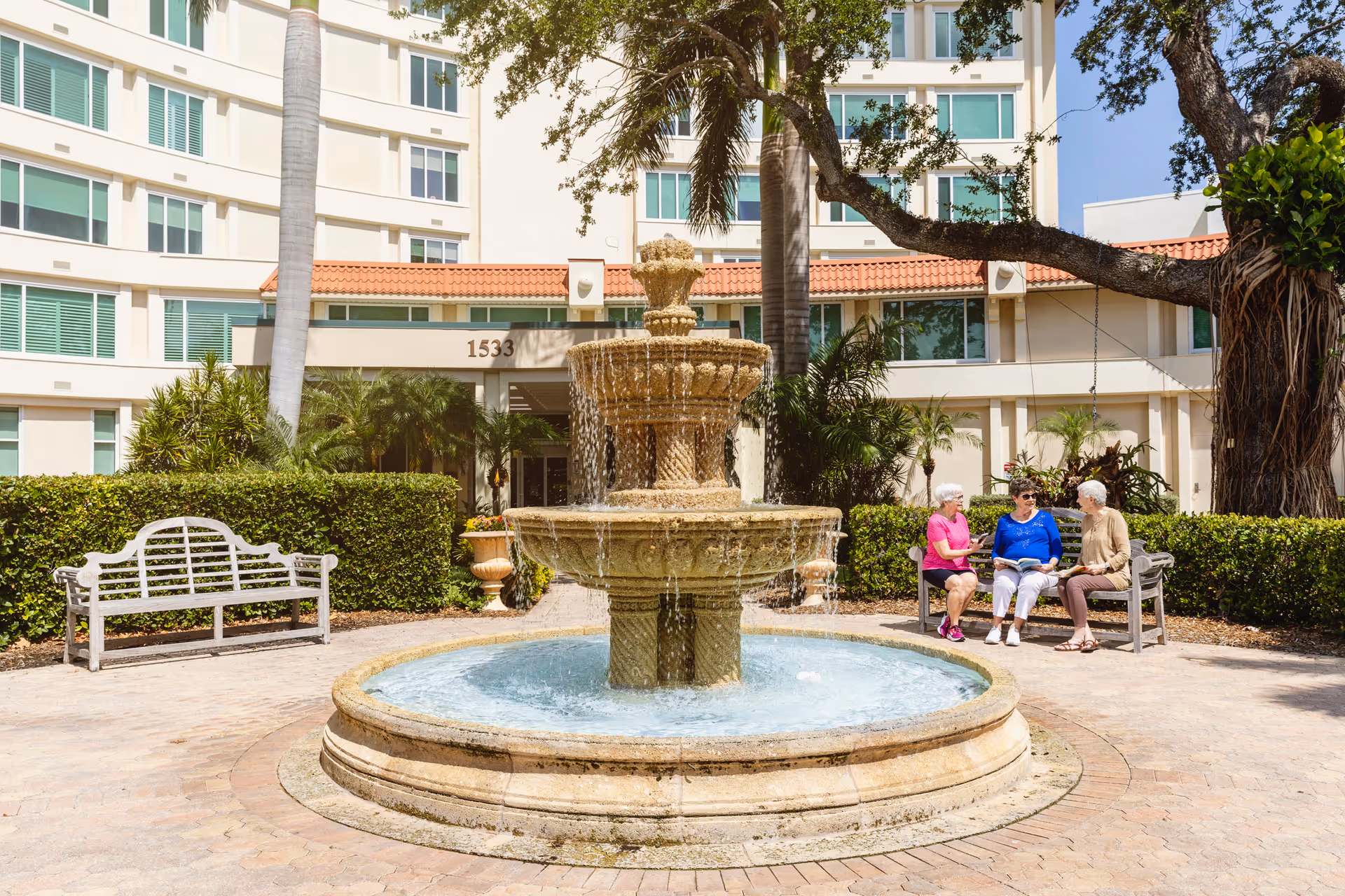 Three elderly women sitting and chatting on benches near a large stone fountain in an outdoor courtyard area of a senior living facility with palm trees and a multi-story building in the background.