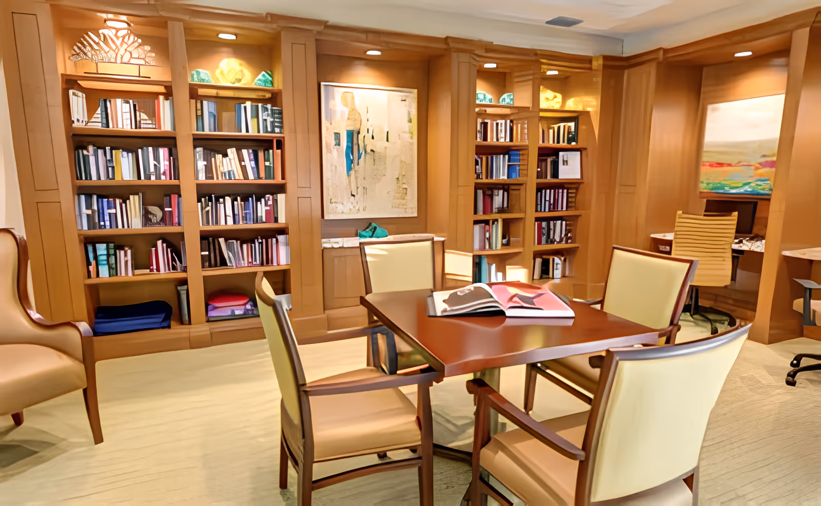 Cozy communal reading room with wooden bookshelves, a central table surrounded by four chairs, and artwork on the walls.