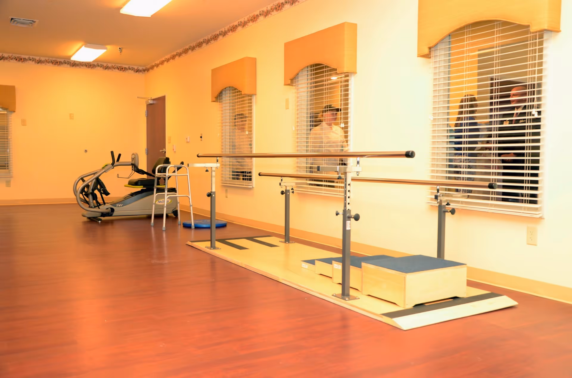 A rehabilitation or physical therapy room with wooden flooring, parallel bars for walking exercises, a walker, an exercise bike, and windows with blinds. There are people visible through the windows in an adjacent room.
