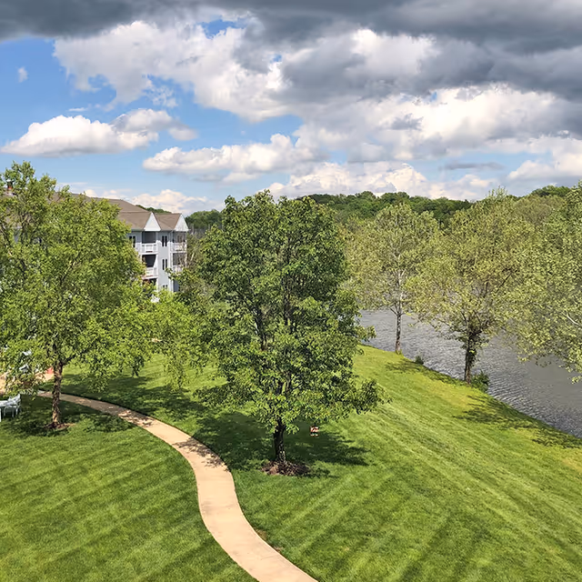 A scenic outdoor view at Cape Albeon Independent Living featuring a winding concrete pathway through a well-maintained grassy area with several green trees. In the background, there is a calm river and a multi-story residential building partially visible on the left side under a partly cloudy blue sky.