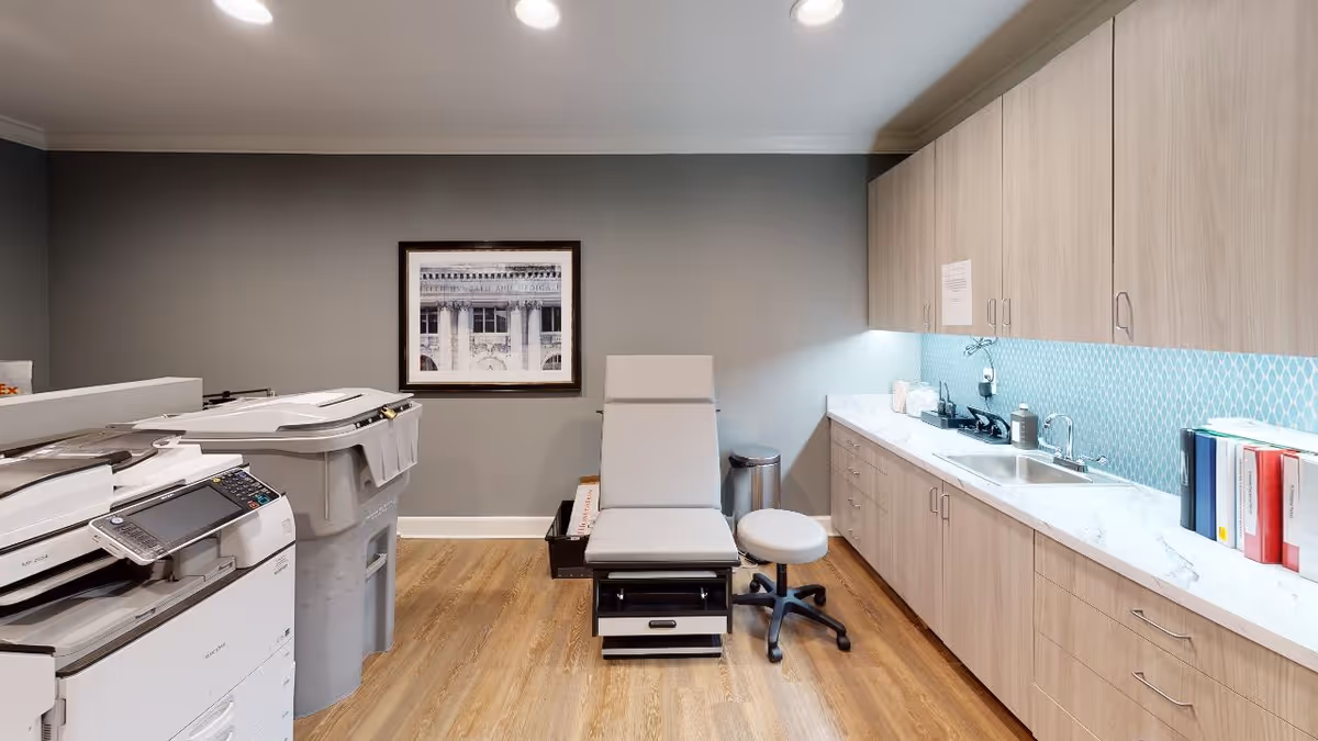 A medical or examination room with a white examination chair and a matching stool in the center. To the right, there is a long countertop with a sink, cabinets above and below, and some books and medical supplies. On the left side, there are two large office machines, likely printers or copiers. The walls are painted gray, and there is a framed black and white photograph hanging on the wall. The floor is wooden.