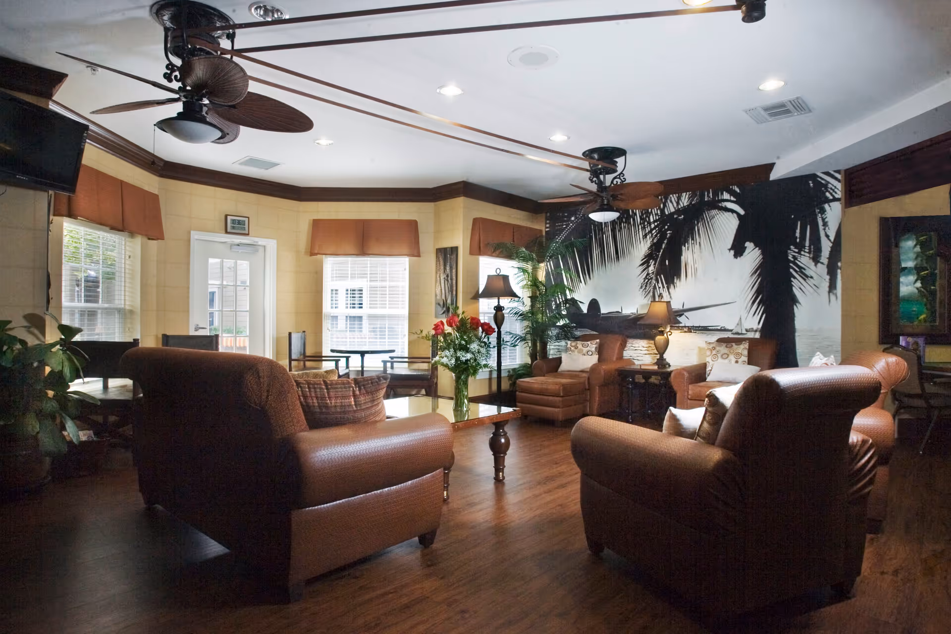 A cozy living room area with brown leather armchairs and a sofa arranged around a wooden coffee table with a vase of flowers. The room features large windows with brown valances, ceiling fans with lights, and a large wall mural of palm trees and an airplane. There are also floor lamps and indoor plants adding to the warm ambiance.