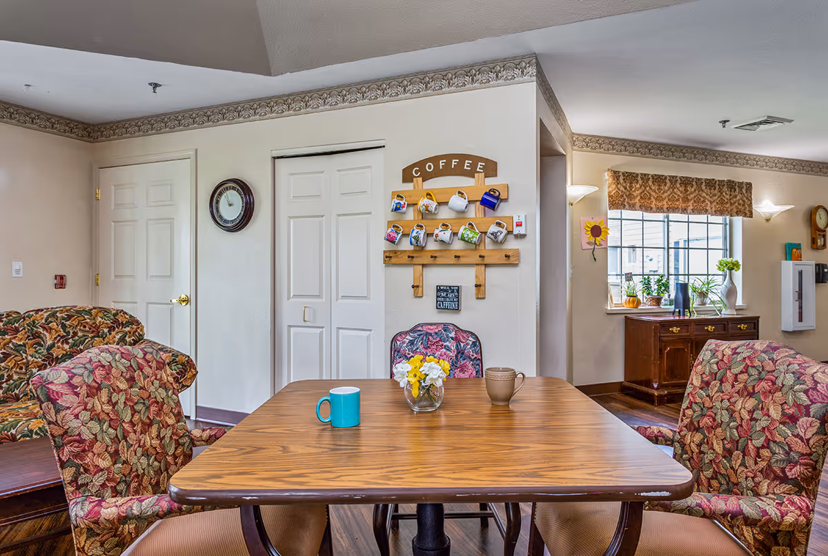 A cozy interior room with a wooden table and four floral upholstered chairs. On the table are two coffee mugs and a small vase with yellow and white flowers. Behind the table, a wall-mounted wooden rack holds colorful coffee mugs under a sign that reads 'COFFEE'. To the right, a window with a patterned valance lets in natural light, illuminating a wooden cabinet with potted plants and a vase. The room has light-colored walls with decorative trim and a clock mounted on the wall.