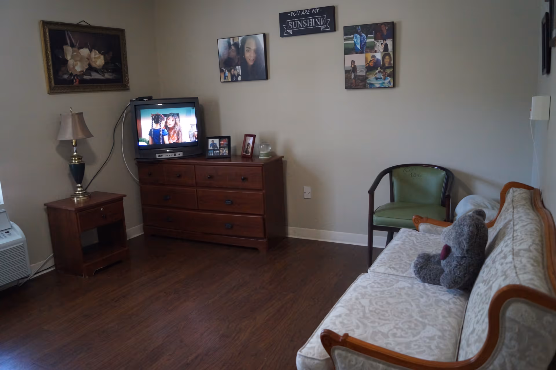 A cozy living room with a wooden floor, a patterned cream sofa with a gray teddy bear on it, a green armchair, a wooden dresser with a small TV showing a scene with two people, a table lamp on a small wooden side table, and several framed photos and paintings on the walls.