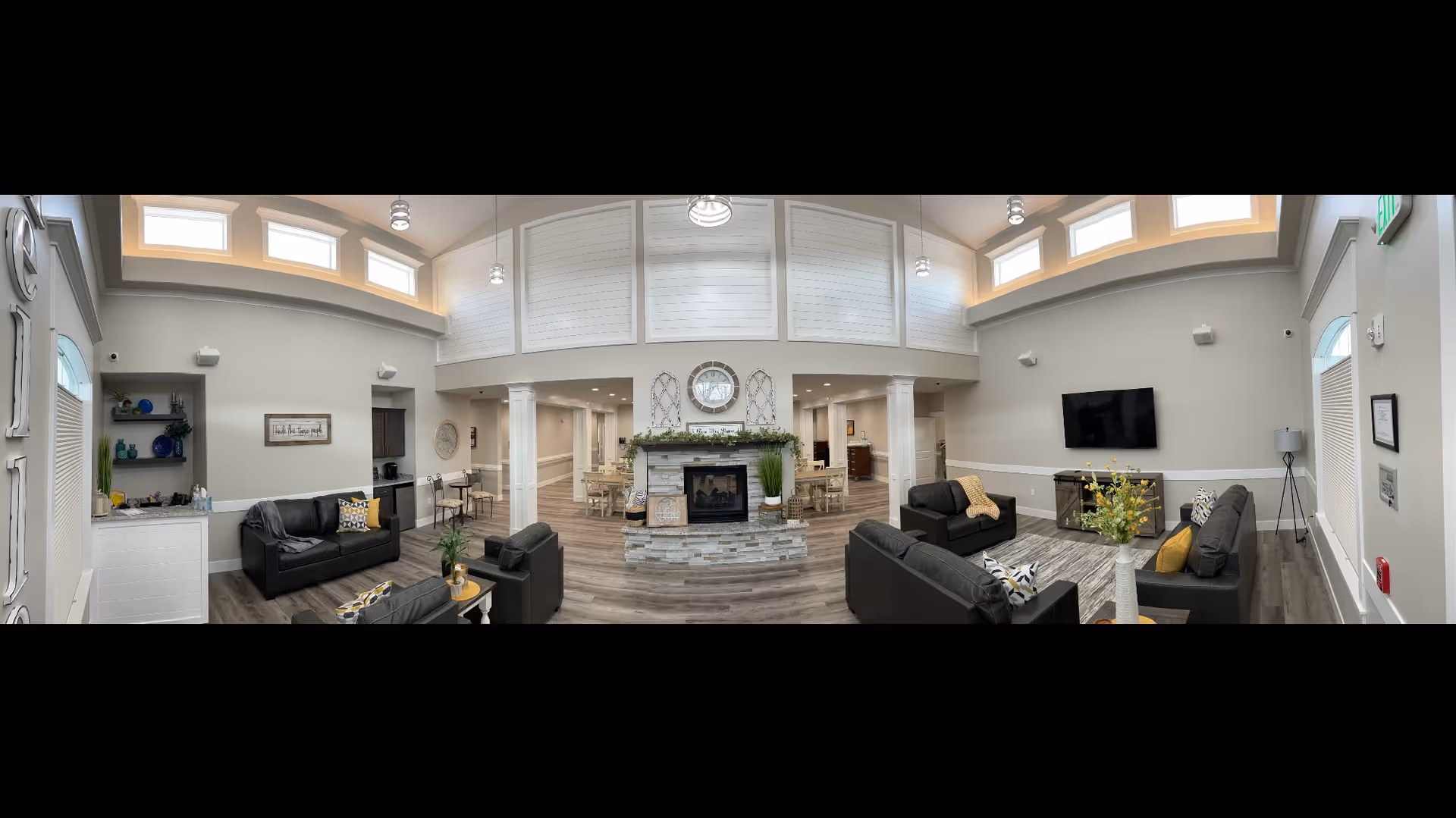 A spacious and well-lit senior living facility common area featuring multiple black leather sofas and armchairs arranged around a central stone fireplace with decorative greenery and a clock above it. The room has high ceilings with small windows near the top, pendant lights, and wood flooring. There is a flat-screen TV mounted on the wall, a small table with a vase of flowers, and dining tables visible in the background. The walls are painted light gray with white trim, and there are decorative shelves and framed artwork on the walls.
