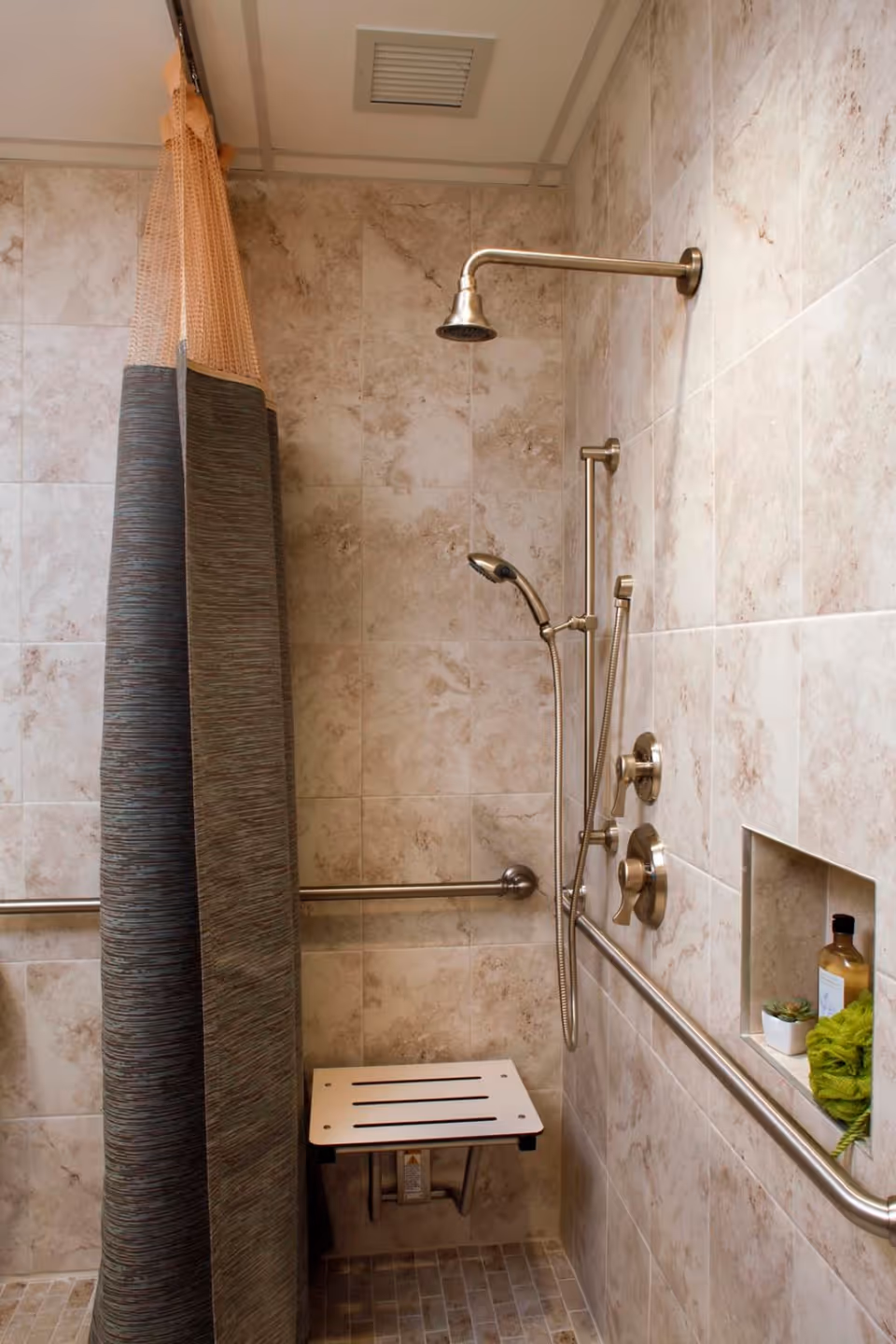 A tiled shower area with a wall-mounted showerhead, handheld shower, grab bars, and a fold-down shower seat. There is a shower curtain partially drawn to the side and a recessed shelf holding a bottle, a small plant, and a green bath sponge.