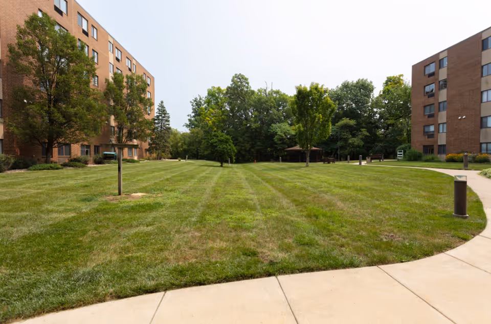 A large grassy courtyard area with a few small trees and a gazebo in the background, surrounded by two multi-story brick buildings with windows. A curved concrete walkway runs along the right side of the grass.