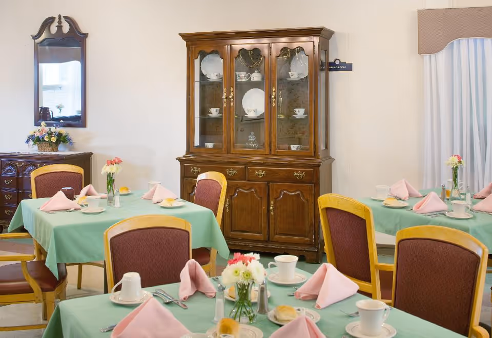 Dining room with tables set with green tablecloths, pink napkins, cups and floral centerpieces and a wooden china cabinet.