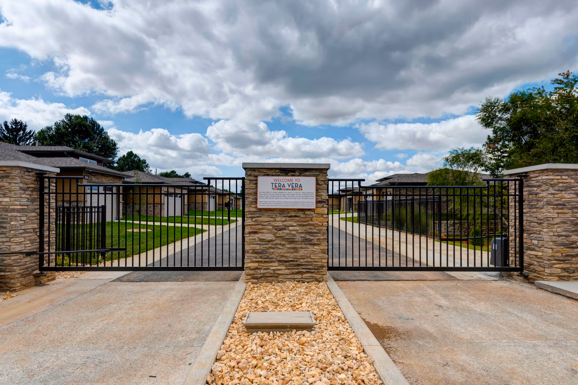 Entrance gate of Tera Vera 55+ facility with a stone pillar in the center displaying a welcome sign. The gate is black metal with a driveway leading into the community, flanked by stone walls and residential buildings visible in the background under a partly cloudy sky.