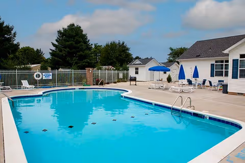 Outdoor swimming pool with clear blue water surrounded by a concrete deck. Several lounge chairs and blue umbrellas are arranged near a white building. Trees and a fence are visible in the background under a partly cloudy sky.