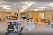 Spacious senior therapy and activity room with exercise equipment, parallel bars, and a kitchenette area under ceiling lights.