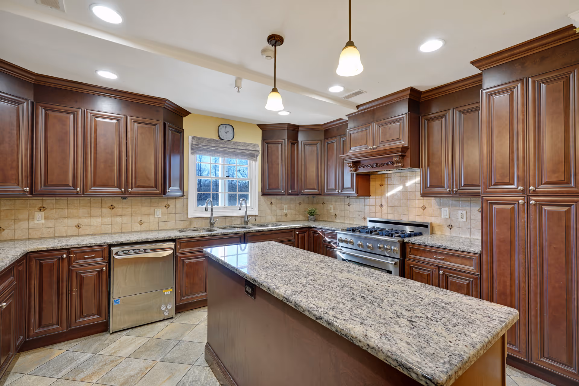 A spacious kitchen with dark wooden cabinets, granite countertops, a central island, stainless steel dishwasher, and a large stove with an ornate wooden hood. There is a window above the sink with a clock mounted above it, and two pendant lights hanging from the ceiling.