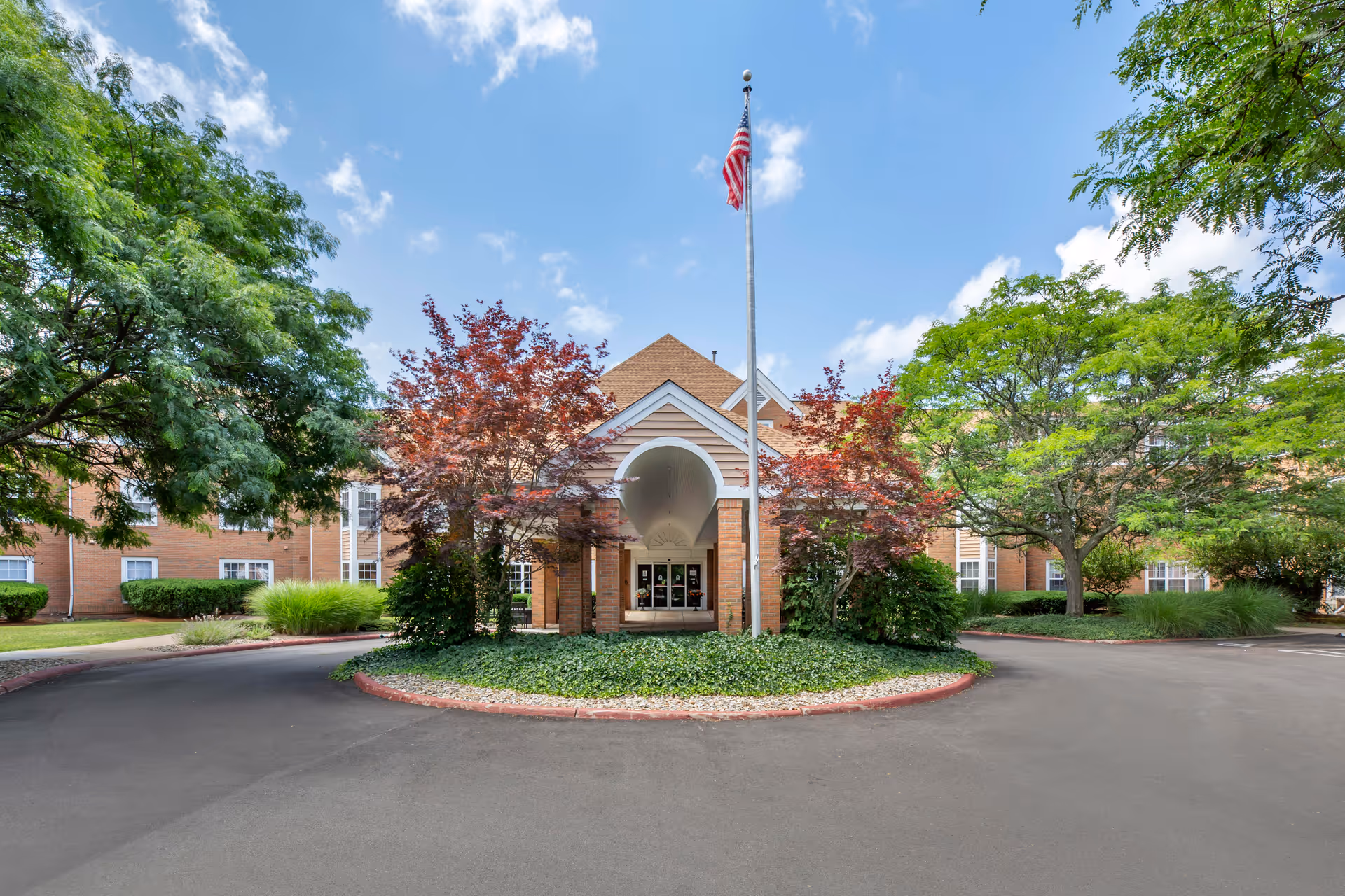 Front exterior view of Brookdale Montrose facility with a circular driveway, landscaped greenery including trees and bushes, and an American flag on a flagpole in front of the entrance under a blue sky with some clouds.