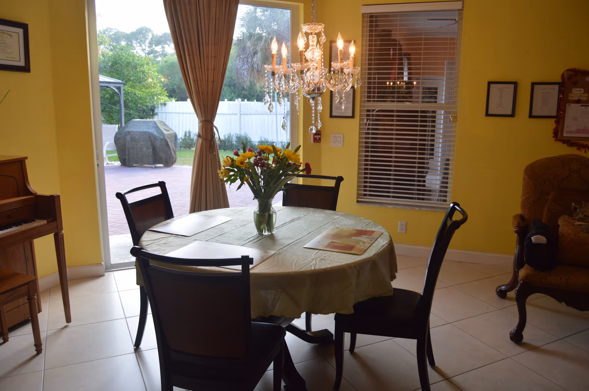 A dining area with a round table covered with a light-colored tablecloth and four chairs around it. A vase with colorful flowers is placed in the center of the table. A chandelier with lit candles hangs above the table. To the left, there is a piano, and in the background, a sliding glass door opens to an outdoor patio with a covered grill. The walls are painted yellow, and there is a window with blinds on the right side.