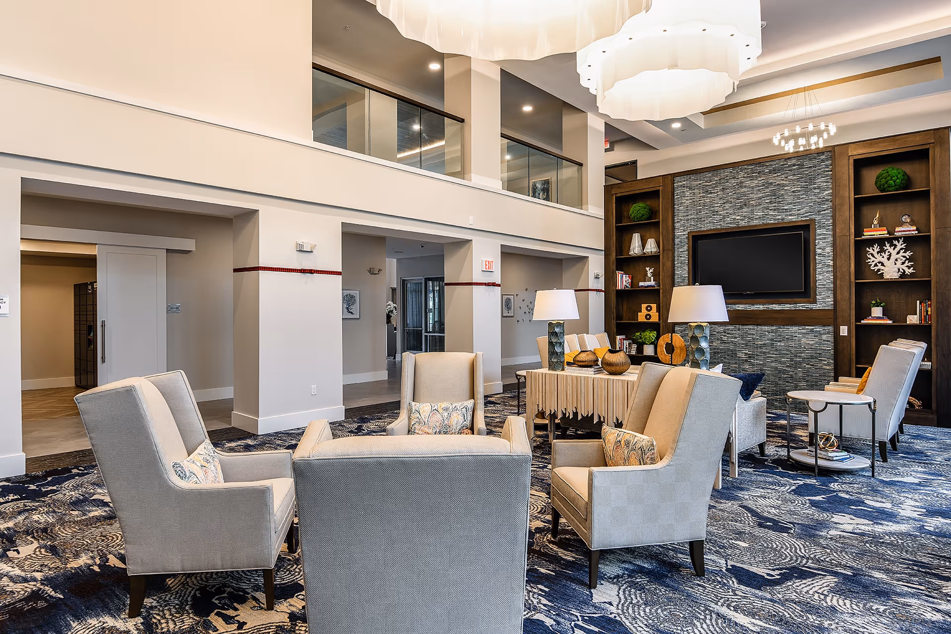 A spacious and elegant living room area with beige armchairs arranged around a central table. The room features a large patterned blue carpet, modern light fixtures hanging from the ceiling, and a wall-mounted flat screen TV surrounded by built-in wooden shelves decorated with books and ornamental items.