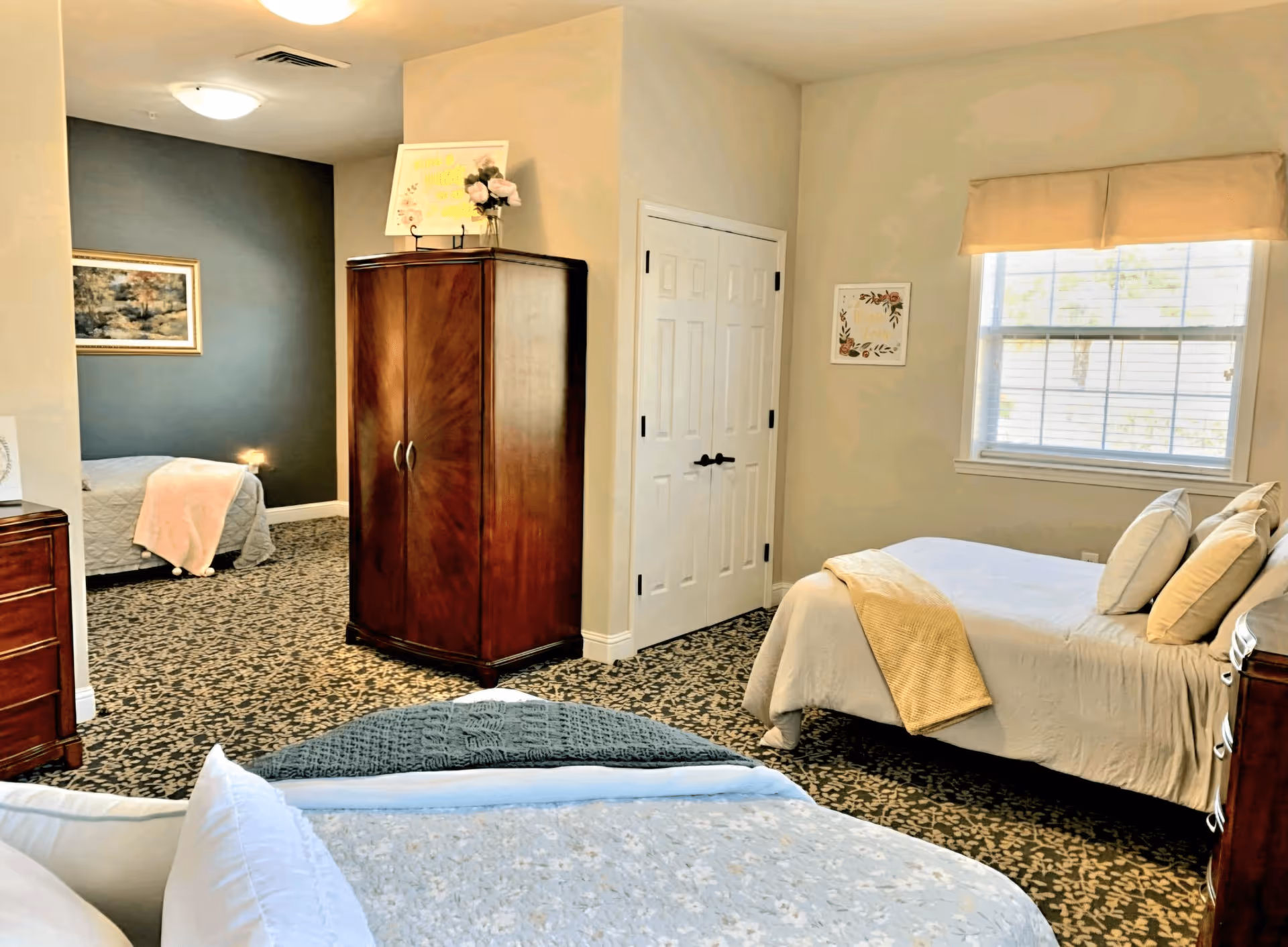 A cozy bedroom area in a senior living facility with two beds, one in the foreground and one near a window with beige curtains. The room features patterned carpet, a wooden armoire, a dresser, and soft lighting. There is a small adjoining space with another bed and a framed picture on a dark accent wall.