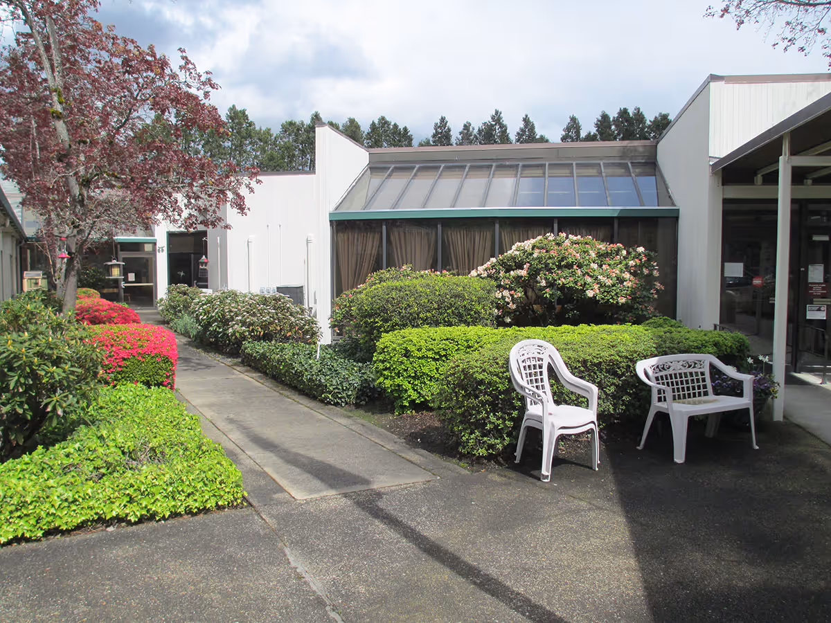 Outdoor courtyard area of a nursing and rehabilitation center with a concrete pathway surrounded by well-maintained bushes and flowering plants. Two white plastic chairs and a white plastic bench are placed on the right side near the building entrance. The building has large windows with curtains and a slanted glass roof section. Trees and a partly cloudy sky are visible in the background.