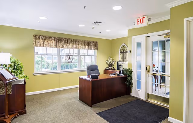 Reception area with a wooden desk and black office chair in front of a large window with a valance. The walls are painted light green, and there is a glass door with an exit sign above it. The room is decorated with plants and a wall ornament.