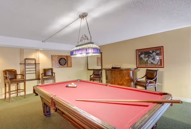 A recreation room with a red-felt pool table, cues, a stained-glass hanging light, chairs, a dartboard, and framed artwork.