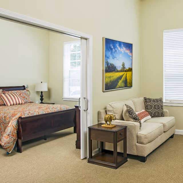 A cozy living area with a beige sofa adorned with patterned cushions next to a wooden side table holding a small elephant figurine. Adjacent to the living area is a bedroom with a wooden bed frame, colorful patterned bedding, a nightstand, and a lamp. Both rooms have windows with white blinds, and a landscape painting hangs on the wall above the sofa.